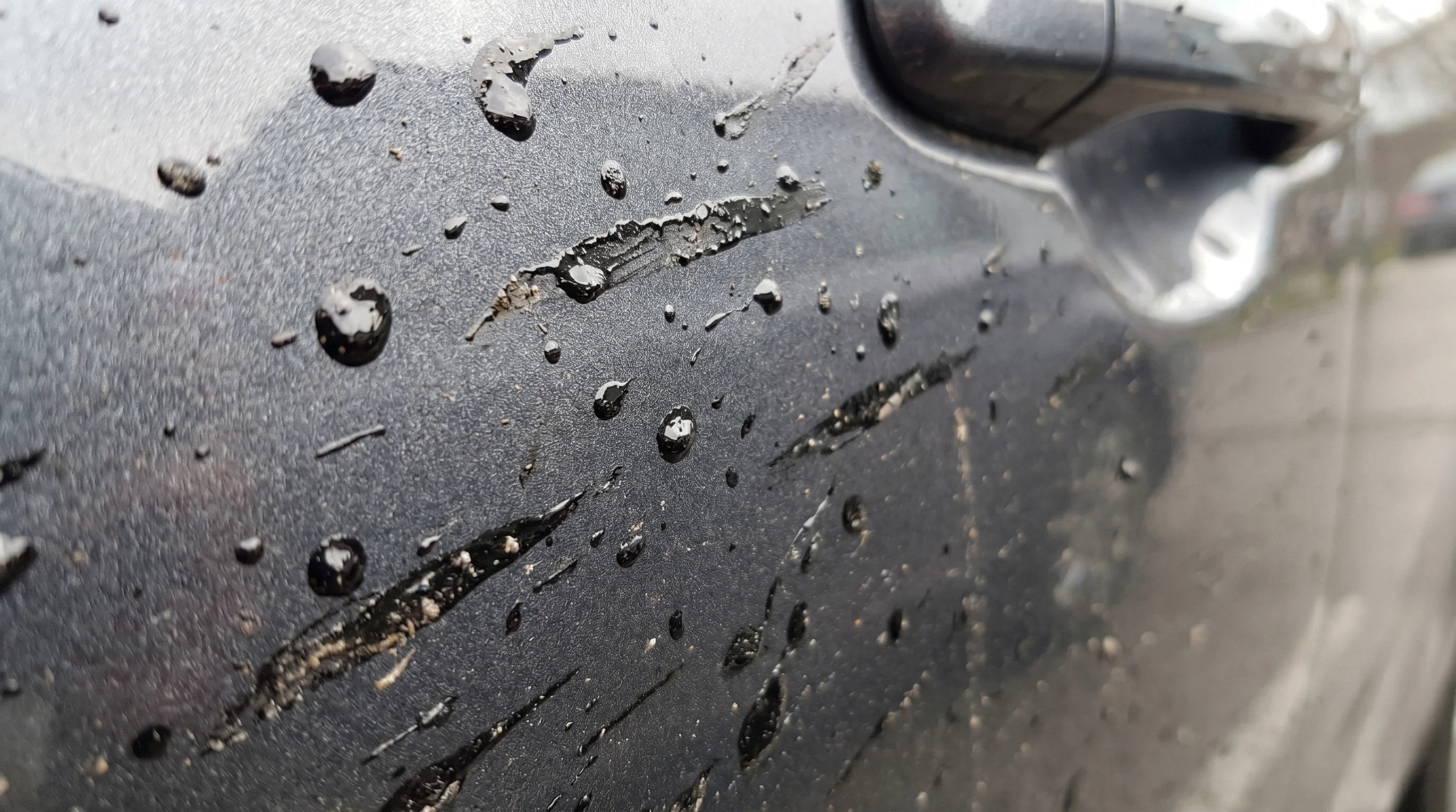 Close-up of dark asphalt and road tar splatter spots on a silver car door panel in bright daylight showing fresh contamination on clearcoat