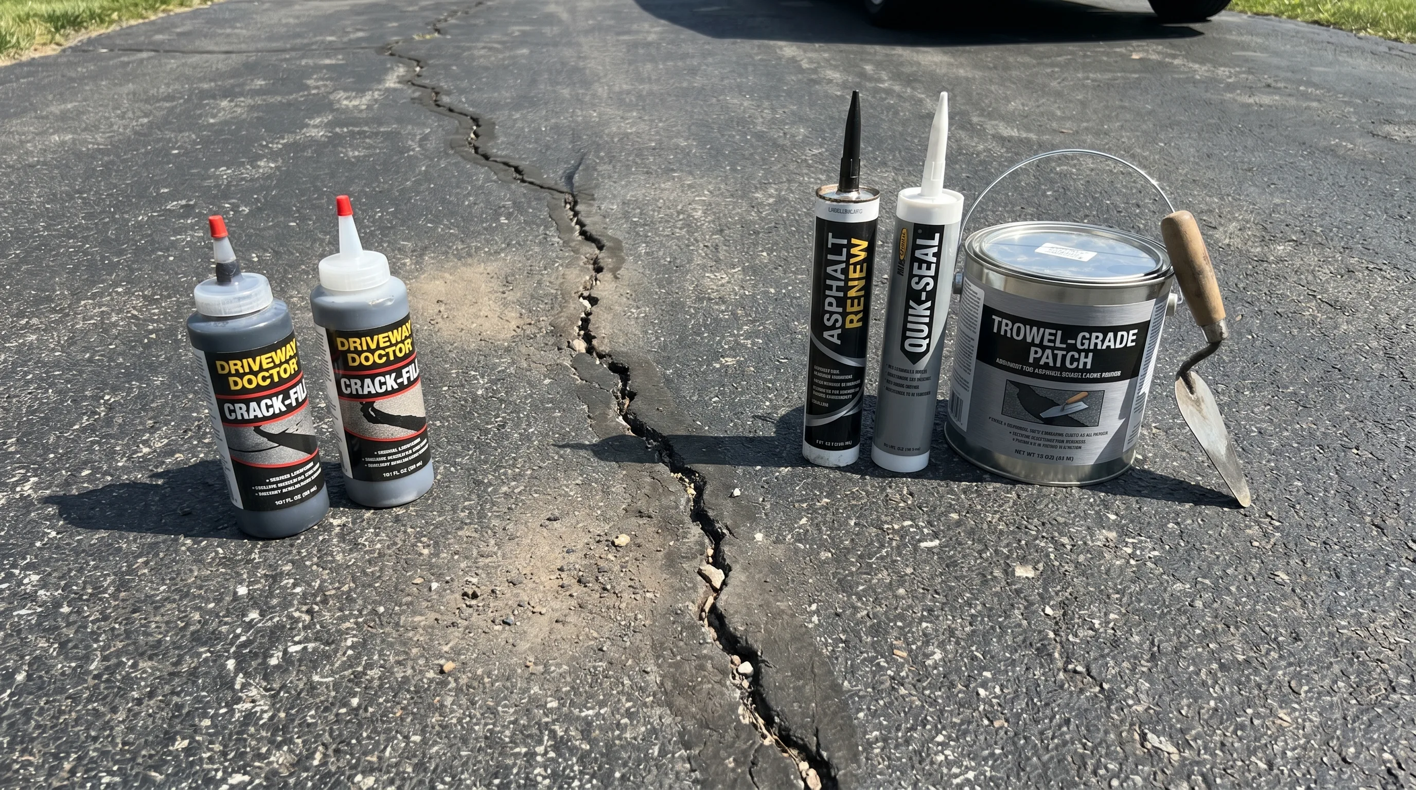Five different asphalt crack filler products — squeeze bottles, caulk tubes, and a trowel-grade pail — arranged on a dark asphalt driveway next to a visible crack in bright daylight